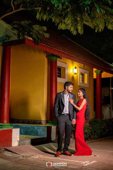 A couple dressed formally stands near a yellow building at night; woman in a red gown touches the man's chest as they gaze at each other.