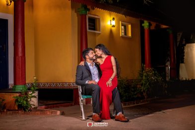 A couple sits close together at night, the woman in a red dress resting her hand on the man's knee, smiling at each other outside a warm-lit building.