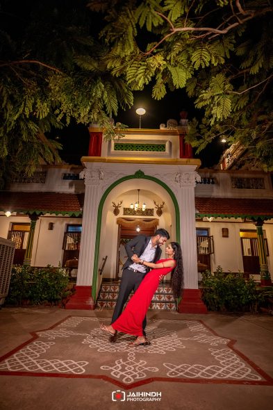 Couple dancing in front of a lit, ornate doorway at night; woman in a red dress and man in a suit, smiling at each other.