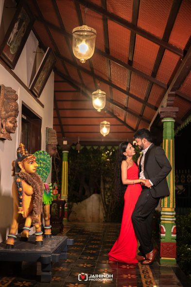 Couple in formal attire embrace and smile in a warm, Indian-themed hallway with statues and ornate pillars at night.",