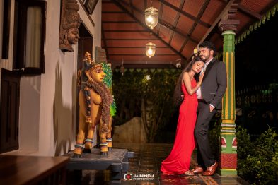 Romantic couple in formal attire posing under a decorated pavilion with green pillar and statue figures at night.