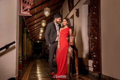 A couple dressed smartly pose in a warmly lit, ornate hallway with carved wooden details, the woman in a red gown and the man in a gray suit.