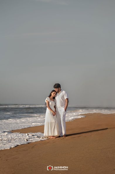 Couple in white outfits standing on a sandy beach, holding hands as waves roll in behind them.