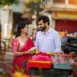 Couple in traditional attire smiles at a flower stall on a busy street, with decorative Tamil text across the top and bottom of the image.