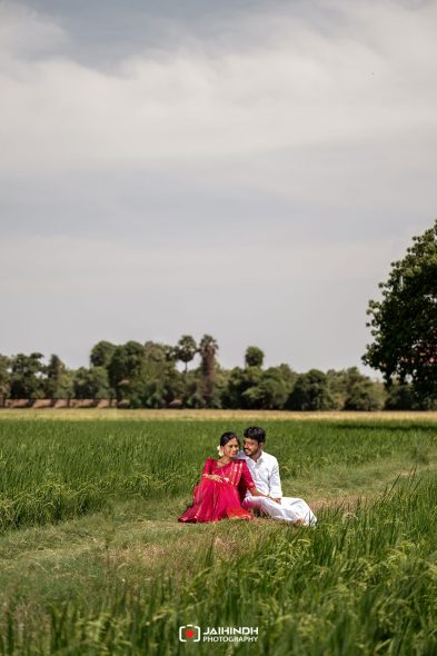Couple sitting close together in a green field, woman in red dress and flower in hair, man in white, with trees in the background under a cloudy sky