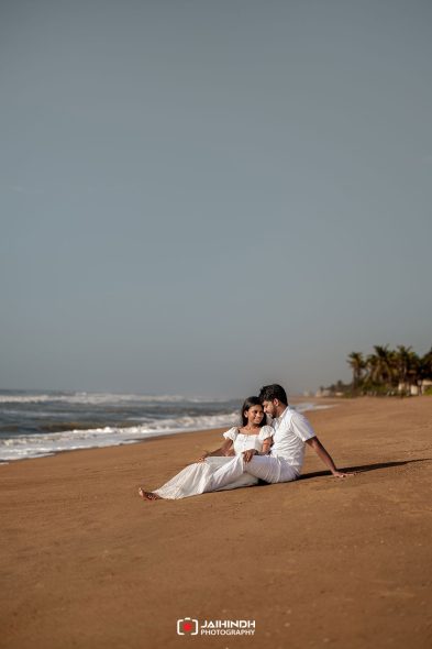 Couple sitting on a sandy beach in white outfits, leaning together and smiling near the shoreline with palm trees in the distance.