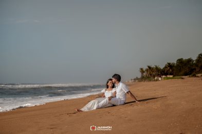 Couple in white sit on the sandy beach as the man kisses the woman on the temple, waves rolling in on the left.
