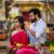 Couple at a flower market; woman in pink traditional dress smiles as man adjusts her hair in the background.