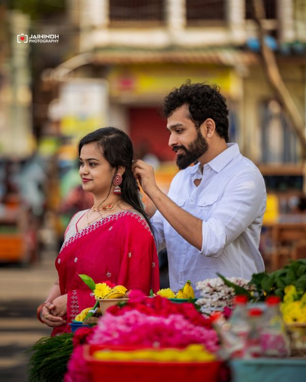 Couple at a flower market; woman in pink traditional dress smiles as man adjusts her hair in the background.