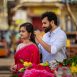 Couple at a flower market; woman in pink traditional dress smiles as man adjusts her hair in the background.