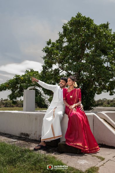 Couple in traditional attire posing by a fountain, the man pointing as they sit on a white wall under a tree.