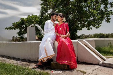A couple sits on a concrete ledge outdoors; the man wears white traditional attire and the woman a pink sari, smiling at each other.