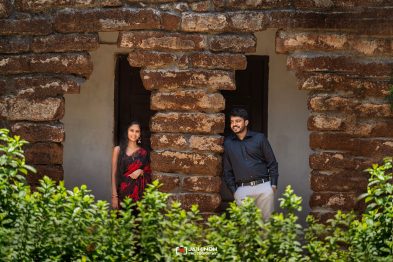 Couple stands beside a weathered stone wall with a central pillar, smiling at the camera; green shrubs in the foreground.