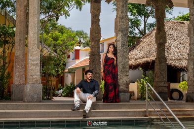Man sitting on steps beside a woman in a red and black saree under stone pillars in a sunny courtyard.