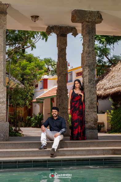 Man seated on poolside steps with a woman in a red and black sari standing beside carved stone pillars under a covered walkway.