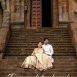 Couple in traditional Indian attire sitting on weathered temple stairs in front of an ornate doorway with carved pillars; wedding scene.