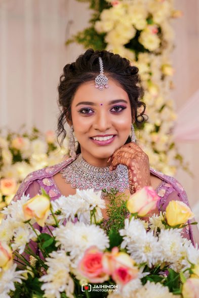 Smiling bride in lavender traditional attire with heavy jewelry, posing among white and pale flowers.