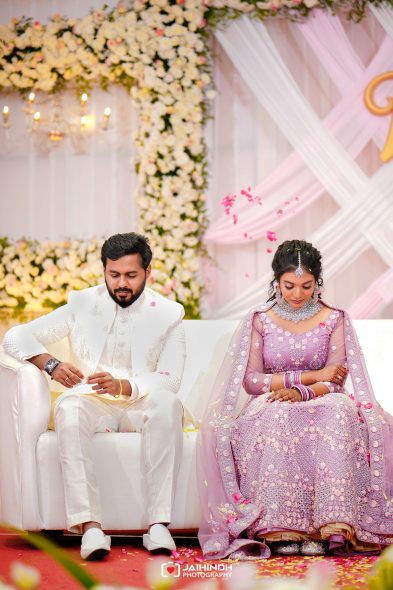 Bride in a pink embellished lehenga and groom in a white suit sit on a white sofa at a wedding ceremony, floral backdrop behind them.