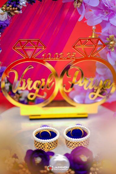Two gold wedding rings rest in pearl-edged holders on a table, with a vivid pink-purple wedding backdrop in the background.