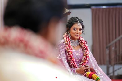 Bride in a lavender-pink traditional outfit wearing elaborate jewelry and flower garlands, smiling at the camera with a blurred guest in the foreground.