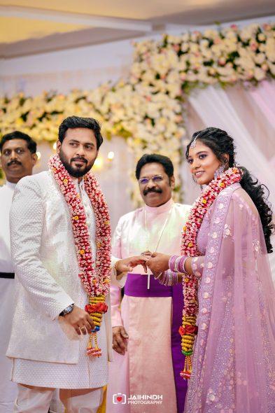Bride and groom in traditional attire exchange rings at a flower-decorated wedding ceremony, wearing garlands around their necks.