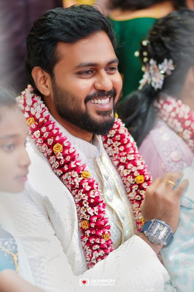 Groom smiling in a white wedding outfit, adorned with pink and white flower garlands at a wedding.
