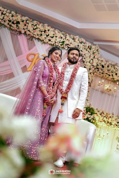 Bride in lavender lehenga and groom in white sherwani pose together at a flower-adorned wedding stage with garlands around their necks.