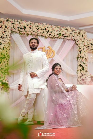 Couple in traditional Indian wedding attire posing on a flower-decorated stage, with the groom standing and the bride seated.