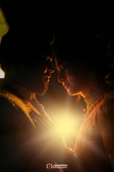 Silhouetted bride and groom lean foreheads together in warm backlight at night, wearing traditional wedding attire and jewelry.