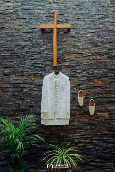 White traditional sherwani with matching footwear hangs under a large wooden cross on a dark stone wall, with green plants in the foreground.