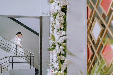 Bride in a white lace wedding dress descending a staircase beside a tall flower wall.