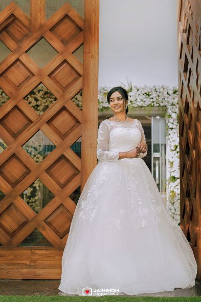 Bride in a white lace wedding gown standing between wooden lattice arches with a floral backdrop, smiling.