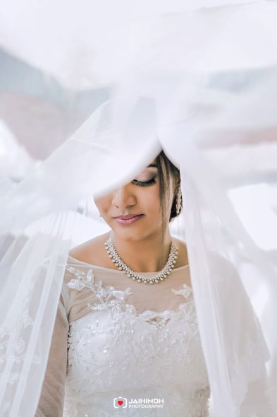 Bride in a lace wedding gown with a pearl necklace, eyes closed under a flowing veil.