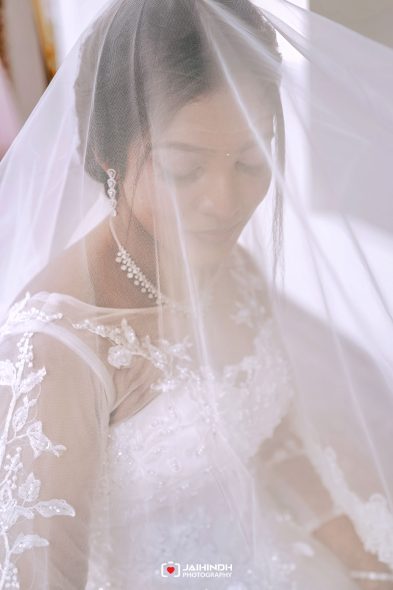Bride in a lace wedding gown, softly veiled, serene expression visible through sheer veil.