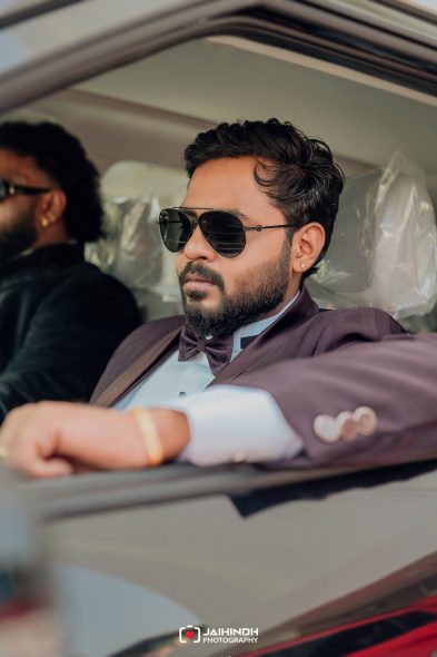 Man in a maroon suit with a bow tie and sunglasses leaning on a car window, looking off to the side.