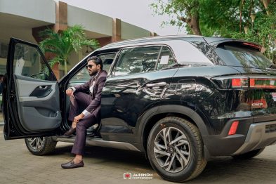 Man in a burgundy suit with sunglasses exits a black SUV with the front door open on a paved driveway.