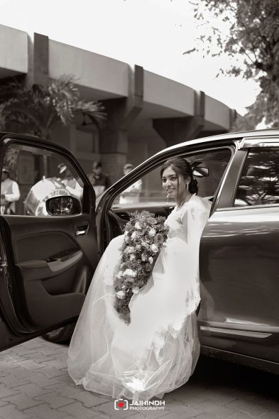 Bride in a wedding gown sits in a car with the door open, holding a cascading bouquet as she prepares to exit the vehicle against an urban backdrop.