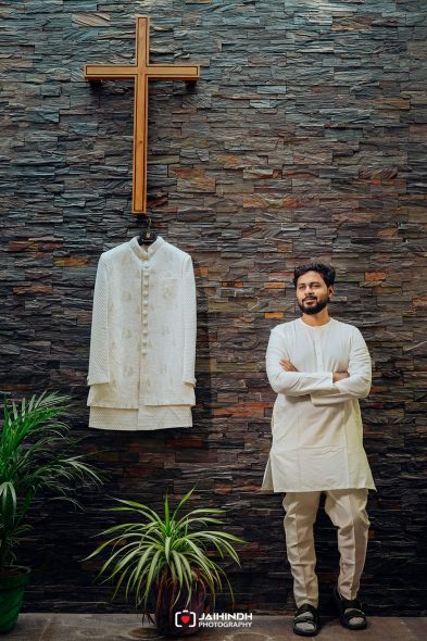 Man in a white kurta posing beside a hanging white sherwani against a dark textured wall with a large wooden cross above.
