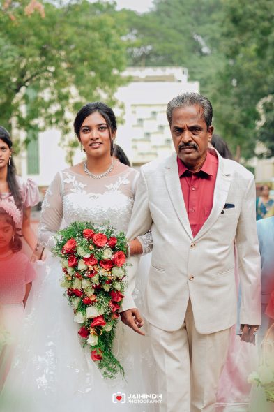 Bride in a white lace wedding gown holding a cascading red-and-white bouquet walks beside an older man in a light suit outdoors, smiling.