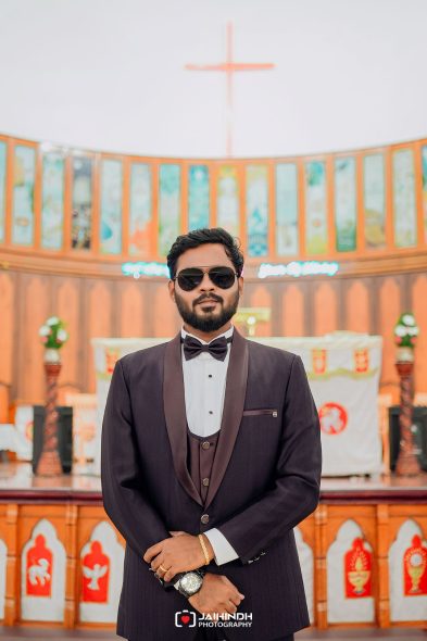 Man in a tuxedo with bow tie and sunglasses posing in front of a church altar with a cross in the background.