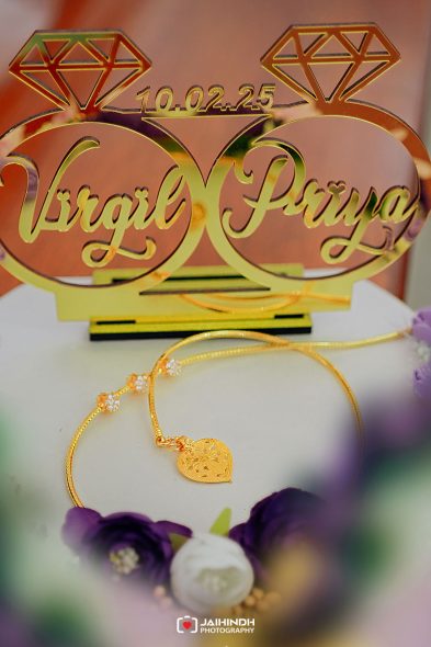 Gold bracelet with heart pendant on a white surface, a blurred floral foreground and a decorative wedding sign in the background showing diamonds and a date.