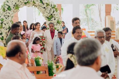 Bride in a white lace gown walking down the aisle under a large white flower arch, escorted by a man in a beige suit, with children and guests nearby.