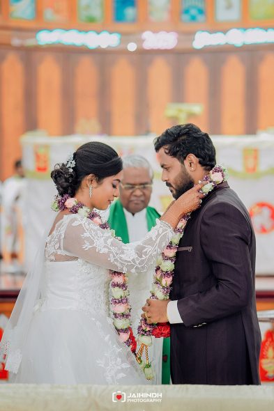 Bride in a white lace gown places a flower garland around groom's neck as they exchange garlands during a wedding ceremony, with a priest in the background.