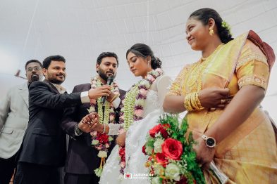 Bride and groom with floral garlands as a man in a suit speaks into a microphone during their wedding ceremony, surrounded by family partners in festive attire.