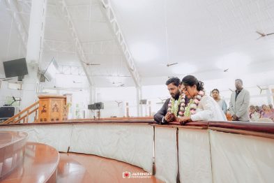 Bride and groom wearing flower garlands bow their heads in prayer at a curved wooden railing during a daytime indoor wedding, guests visible in the background.]','Bride and groom clasp hands and pray at a curved railing, garlands around their necks in a bright indoor ceremony with family nearby.]','Indoors wedding scene: couple in the foreground pray at a rounded wooden barrier, floral garlands adorning them as guests stand behind.]','A couple at a wedding lean on a curved railing, hands joined in prayer, surrounded by family in a spacious hall.]','Wedding ceremony inside a bright hall; couple in the foreground with garlands prays at the railing while guests observe.]