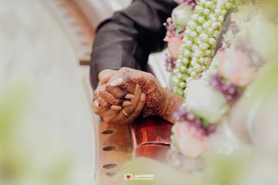 Close-up of a bride and groom holding hands, with intricate henna designs and a floral garland in the background.