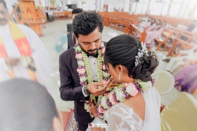 Bride and groom exchange flower garlands during a wedding ceremony indoors, with guests seated in the background.
