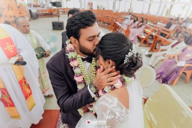Bride and groom kiss during their wedding ceremony, adorned with flower garlands, in a church with guests watching.