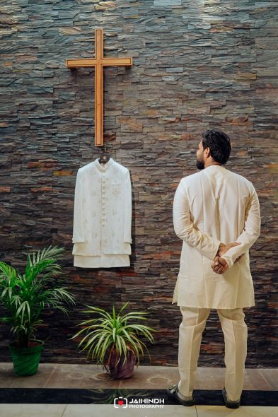 Back view of a man in cream traditional clothing standing with his hands behind his back, facing a wooden cross on a stone wall with a white garment hanging nearby."