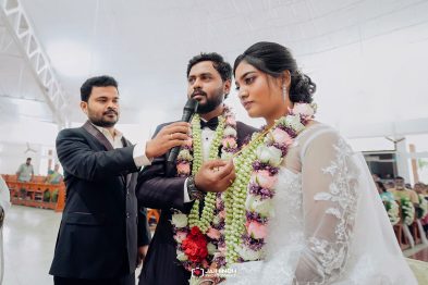 Bride and groom with floral garlands as a man passes a microphone at their wedding ceremony indoors.